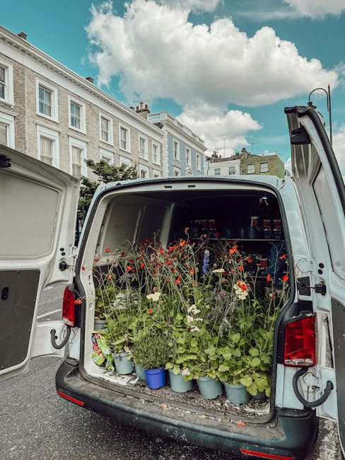 The open rear of a white delivery van filled with potted flowers, including red poppies, white daisies, and green foliage, arranged in light blue and plain pots. The plants are situated on a shelf wit