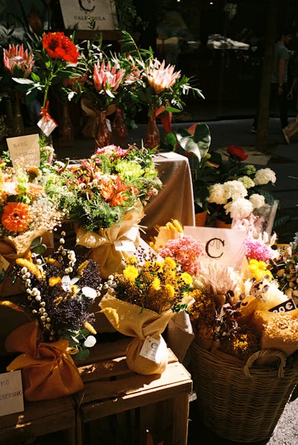 A vibrant display of floral arrangements featuring a variety of fresh flowers, including pink lilies with elongated petals, bright red roses, yellow chrysanthemums, purple and white waxflowers, and wh