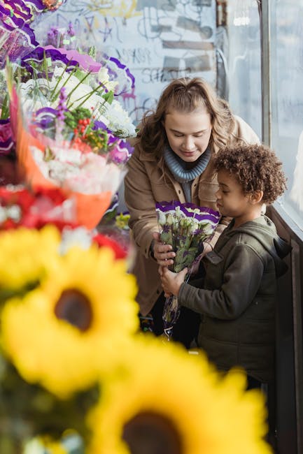 A young woman with wavy blonde hair, dressed in a beige coat and blue turtleneck, presents a small bouquet of purple and white flowers to a young boy with curly brown hair, wearing a dark jacket, insi